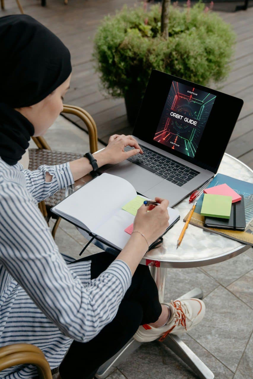 Person sitting at a table outdoors using a laptop with digital AI course 'Orbith Guide' with a notebook and stationery items.
